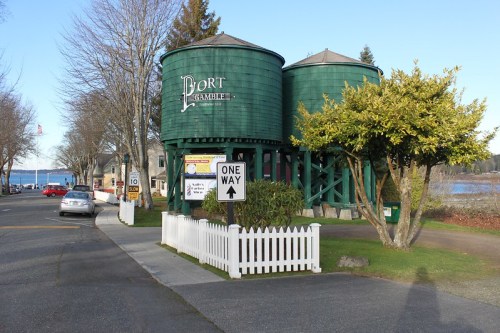 Two green water towers labeled "Port Gamble" beside a road, with a white picket fence and trees in the foreground.