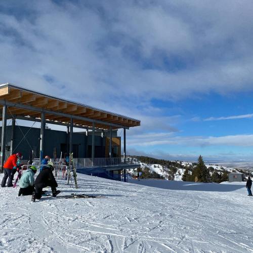 A modern ski lodge on a snowy slope, with people preparing to ski under a blue sky with scattered clouds.