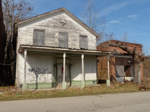 Abandoned two-story building with boarded windows and graffiti, surrounded by overgrown vegetation and a nearby brick structure.