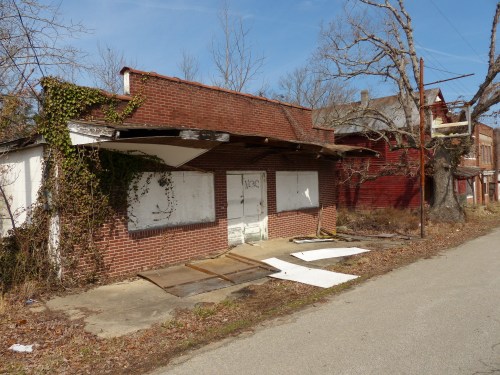 Abandoned brick building with boarded windows, overgrown vines, and debris on a quiet street.