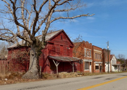 Abandoned red barn and old brick buildings along a quiet road, with a large tree and clear blue sky in the background.