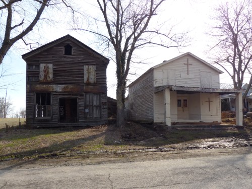 Two old, weathered buildings stand side by side, one wooden and dilapidated, the other painted white with crosses.