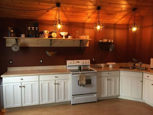 A cozy kitchen with white cabinets, a stove, wooden ceiling, and decorative shelves with dishes and hanging lights.