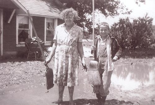 A woman and a boy stand in a muddy area, holding a broom and a dustpan, in front of a house.