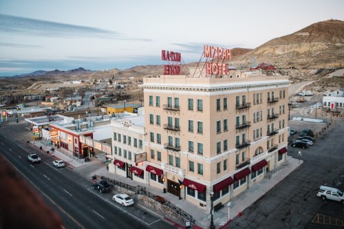 Historic hotel with neon signs, surrounded by a small town and mountains in the background, under a clear sky.