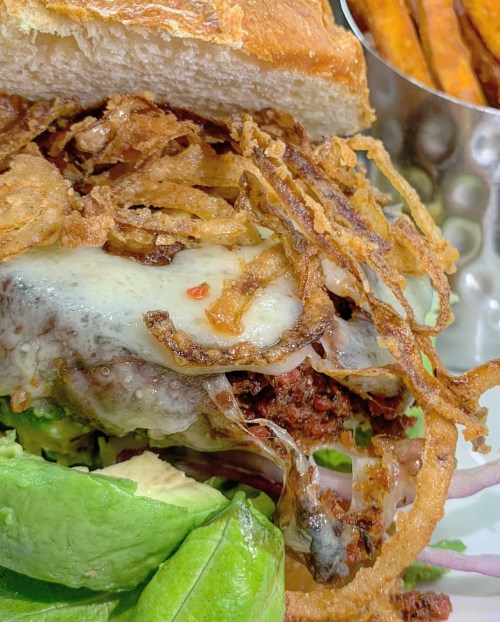 Close-up of a burger with melted cheese, crispy onions, lettuce, and avocado, served with sweet potato fries.