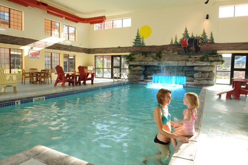 A woman and a young girl play in an indoor pool with a stone waterfall and colorful mural in the background.