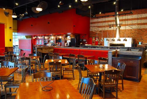 Interior of a restaurant featuring wooden tables, black chairs, a bar area, and a brick oven in a vibrant setting.