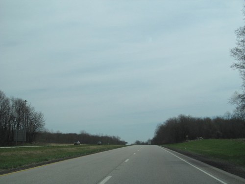A wide, empty highway stretches into the distance under a cloudy sky, flanked by bare trees and grassy areas.