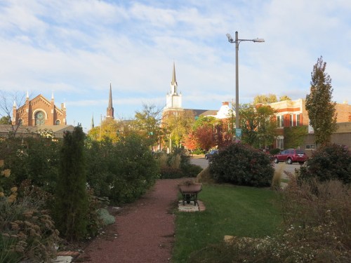 A scenic view of a town with church steeples, trees, and a garden path under a partly cloudy sky.