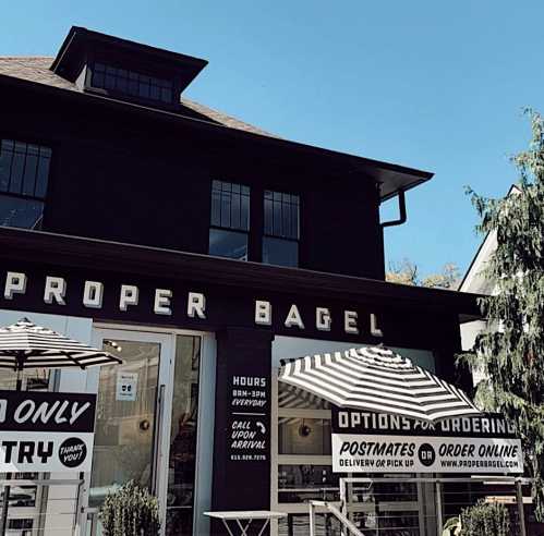 A black building with "Proper Bagel" signage, featuring outdoor seating and signs for ordering options.