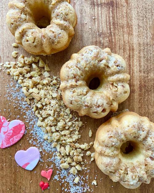 Three small, decorative bundt cakes on a wooden surface, surrounded by crushed nuts and colorful heart-shaped sprinkles.