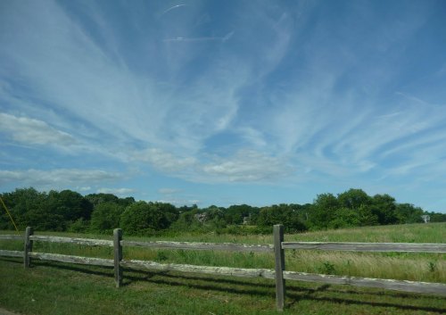 A scenic view of a green field with a wooden fence under a blue sky with wispy clouds.