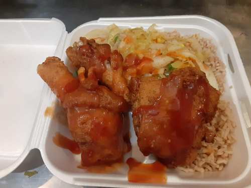 A takeout container with two pieces of fried chicken, drizzled with sauce, served with rice and mixed vegetables.