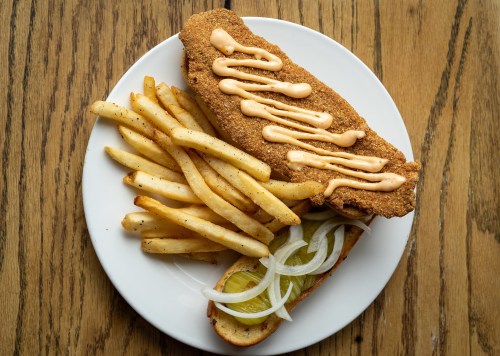 A plate with crispy fried fish, drizzled with sauce, served with French fries and a pickle topped with onions.