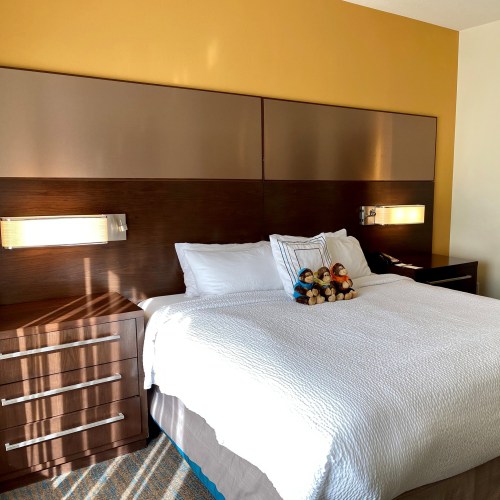 A cozy hotel room with a neatly made bed, bedside lamps, and a wooden dresser. Plush toys are arranged on the bed.