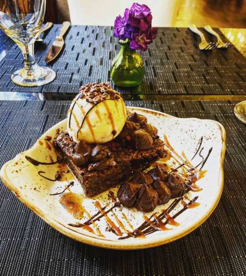 A plate with a chocolate brownie topped with vanilla ice cream, drizzled with chocolate sauce, and a small flower vase.