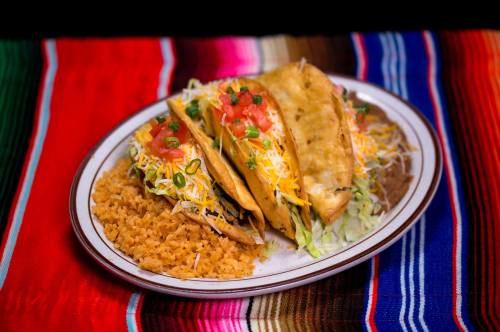 A plate of three tacos topped with lettuce, tomatoes, and cheese, served with a side of rice on a colorful woven tablecloth.