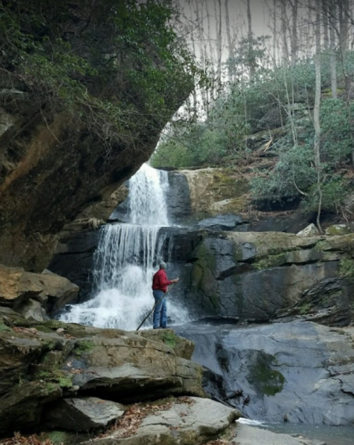 A person in a red jacket stands by a waterfall surrounded by rocky terrain and trees.