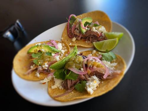 Three tacos on a plate, topped with shredded meat, onions, cilantro, cheese, and lime wedges.