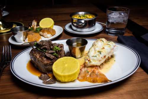 A gourmet meal featuring steak, lobster, lemon, and sides, served on a wooden table with a glass of water.