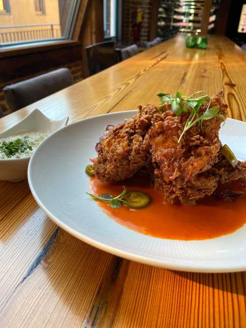 A plate of crispy fried chicken garnished with herbs, served with sliced jalapeños and a side of sauce on a wooden table.