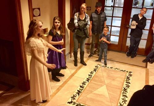 A performer in a dress gestures while a small audience watches in a lobby with decorative flooring.