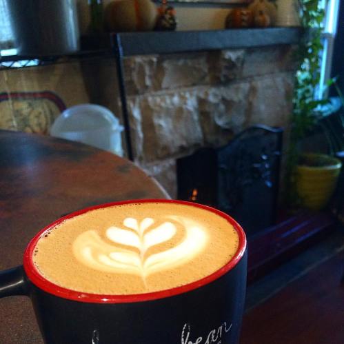 A close-up of a latte in a red mug, featuring a beautiful leaf design in the foam, with a cozy fireplace in the background.