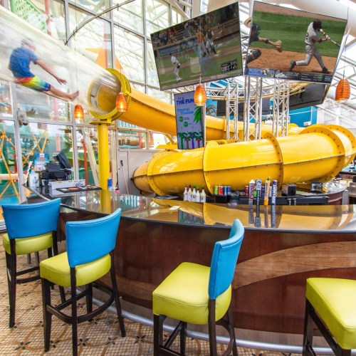 A vibrant bar area with colorful chairs, featuring a large water slide and screens showing sports in the background.