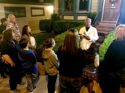 A group of people listens to a speaker in front of a house at night, surrounded by greenery.