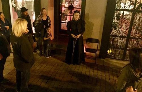 A group of people gathers around a woman in a black dress, standing in a dimly lit street at night.