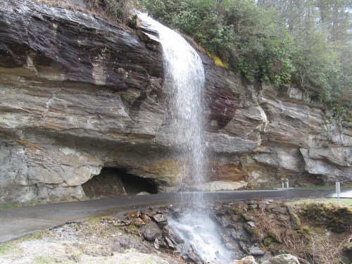 A waterfall cascading down a rocky cliff beside a road, surrounded by greenery and natural rock formations.