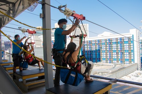 Two people prepare to zip line from a platform, while an instructor assists them in a sunny urban setting.