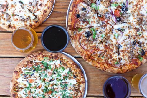 A variety of pizzas on wooden tables, surrounded by glasses of different beverages.