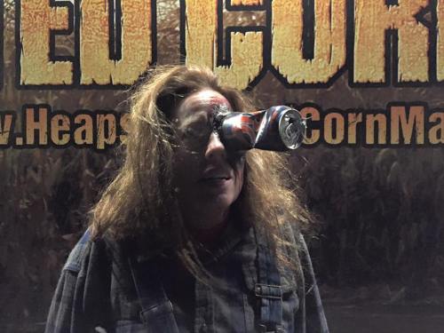 A person with wild hair humorously balancing a soda can on their forehead in front of a corn maze backdrop.
