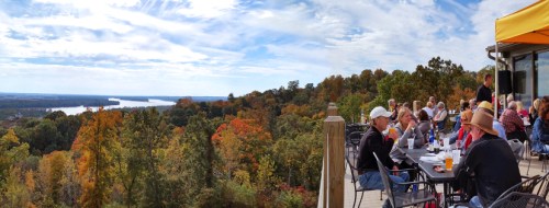 A scenic view from a terrace overlooking a river, with people enjoying drinks and food amidst colorful autumn foliage.