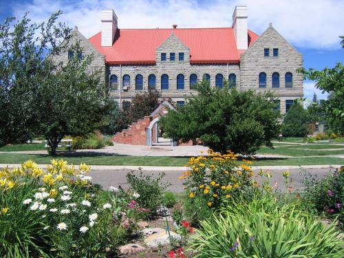 Historic stone building with a red roof, surrounded by colorful flowers and greenery under a blue sky.