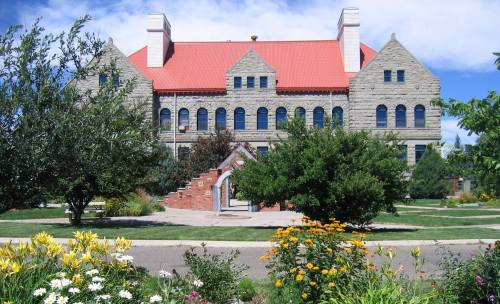 Historic stone building with a red roof, surrounded by colorful flowers and greenery under a blue sky.