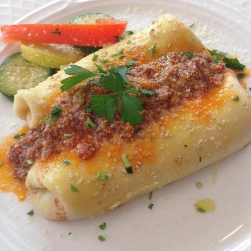 A plate of rolled pasta topped with meat sauce, cheese, and garnished with herbs, served with sliced vegetables.