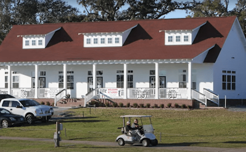 A white building with a red roof, featuring a porch, surrounded by grass and a golf cart in the foreground.