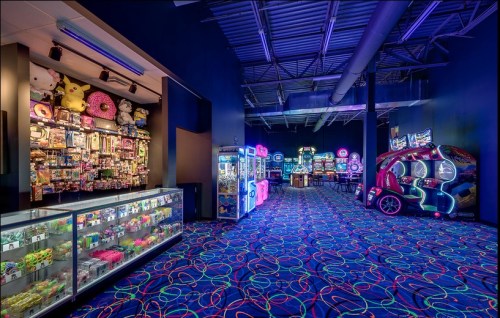 A vibrant arcade with colorful carpet, glowing games, and a display of plush toys and prizes along the walls.