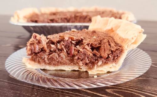A slice of chocolate pie on a clear plate, with a whole pie in the background, resting on a wooden table.