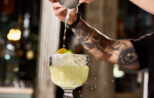 A bartender pours a drink into a glass filled with ice and garnished with lime, with splashes of liquid.