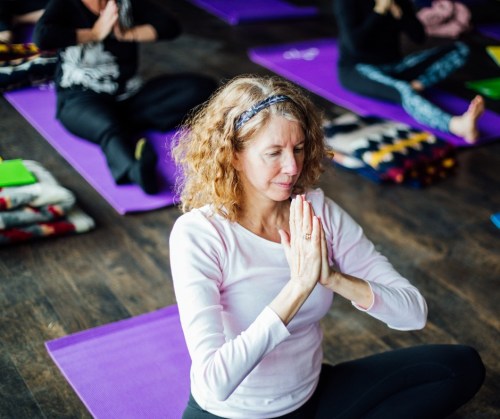 A woman in a pink top sits in a yoga pose with hands in prayer, surrounded by others on yoga mats.
