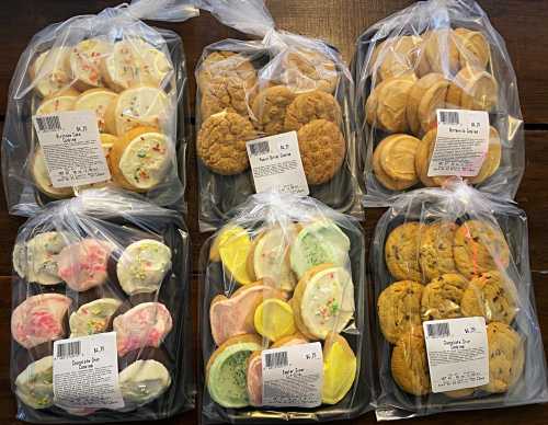 Six trays of assorted cookies, each packaged in clear bags with labels, featuring various colors and shapes.