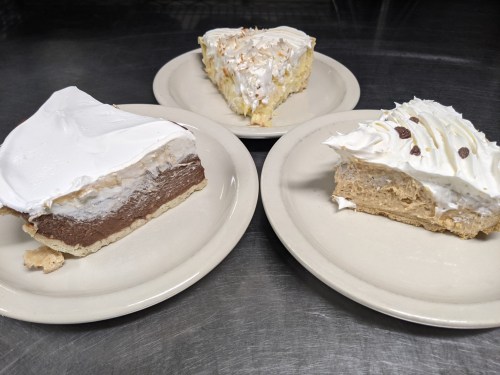 Three slices of pie on plates: chocolate, coconut cream, and a light-colored pie topped with whipped cream.