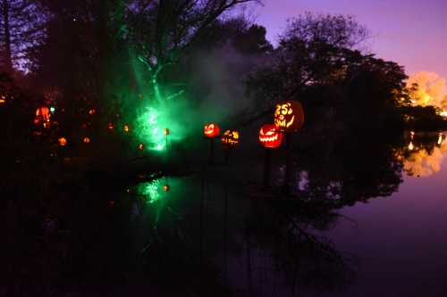 Glowing jack-o'-lanterns reflect on a dark lake, surrounded by mist and eerie green lights at dusk.