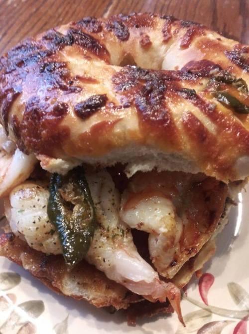 A toasted bagel filled with shrimp, jalapeños, and spices, served on a floral plate.