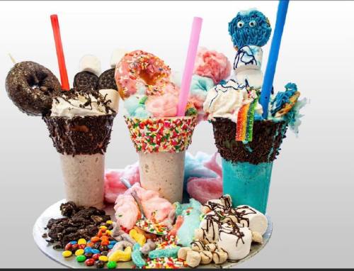 Three colorful milkshakes topped with whipped cream, sprinkles, donuts, and cotton candy, surrounded by candies.