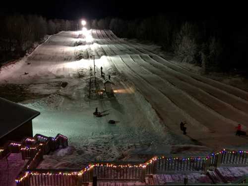 A snowy tubing hill at night, illuminated by lights, with people enjoying sledding down the slopes.